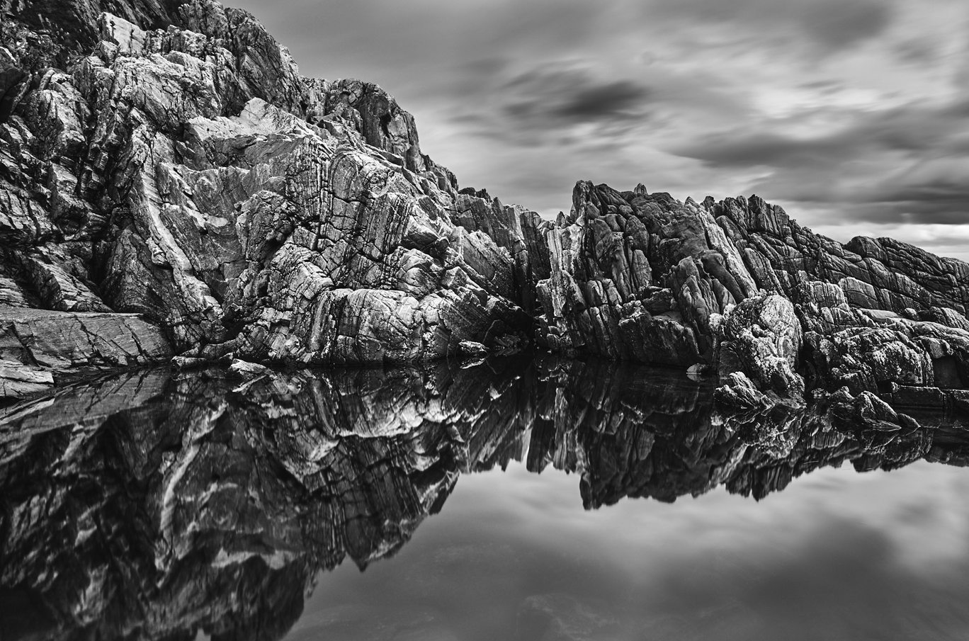 Svarthvitt bilde av solide, taggete klippeformasjoner som speiler seg klart i en stille vannoverflate.