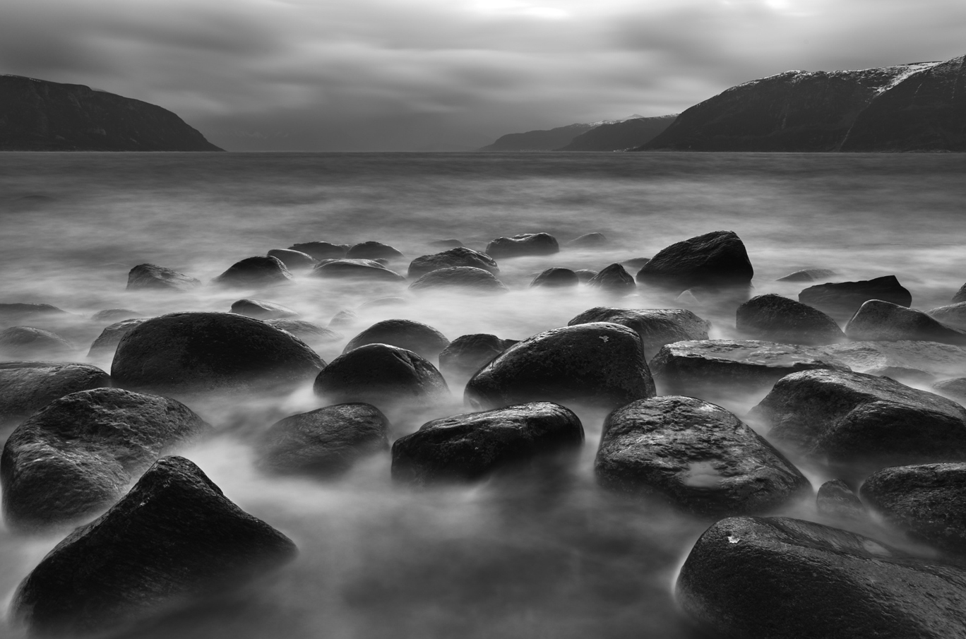 Svart-hvitt bilde av glatte steiner ved en stille fjord med fjell i bakgrunnen