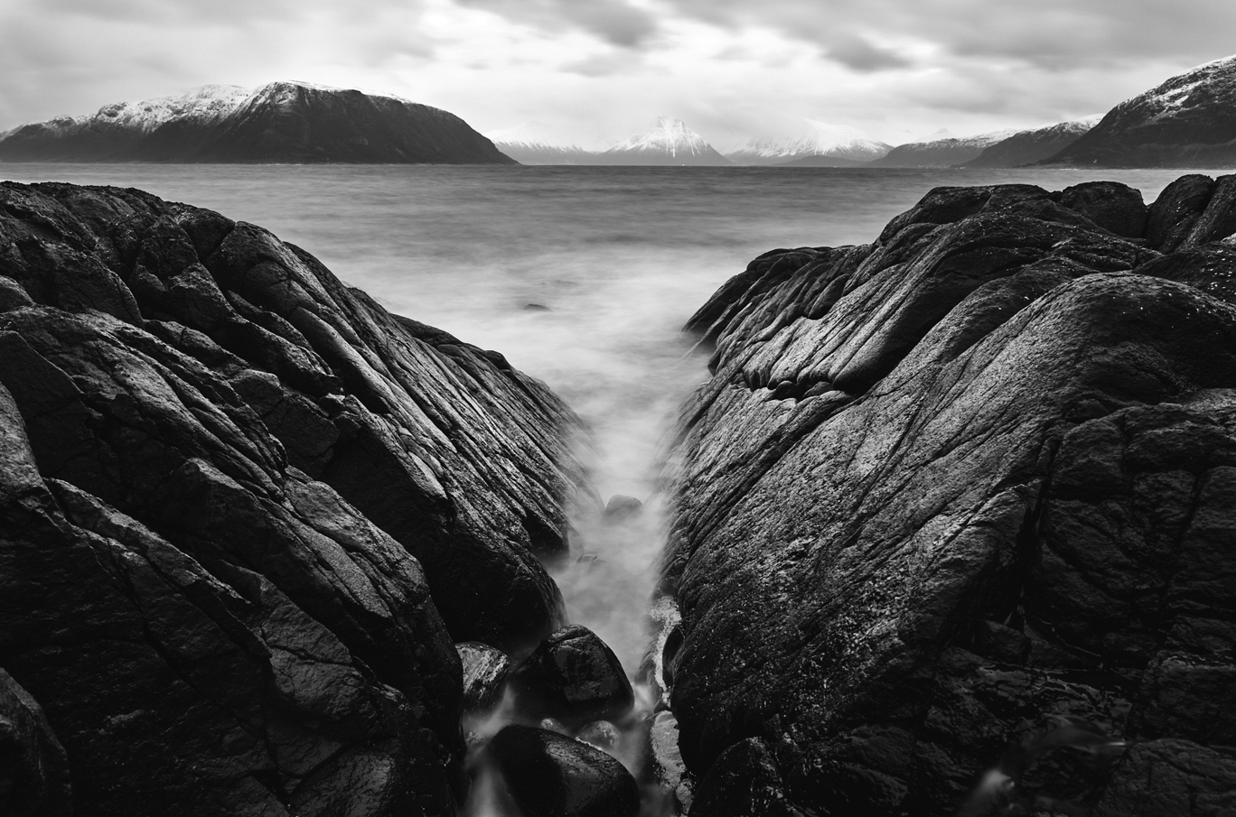 Svarthvitt bilde av steinformasjoner som danner en smal kløft, med havet som strømmer imellom og snødekte fjell i bakgrunnen