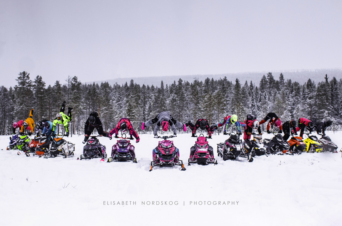 En rekke personer i fargerike klær gjør håndstående på snøscootere i et snødekt landskap med skog i bakgrunnen