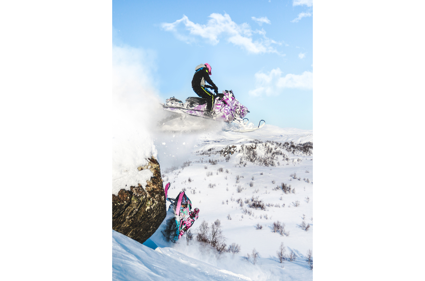 Person som kjører snøscooter i lufta over en snødekt fjellside under en klar, blå himmel