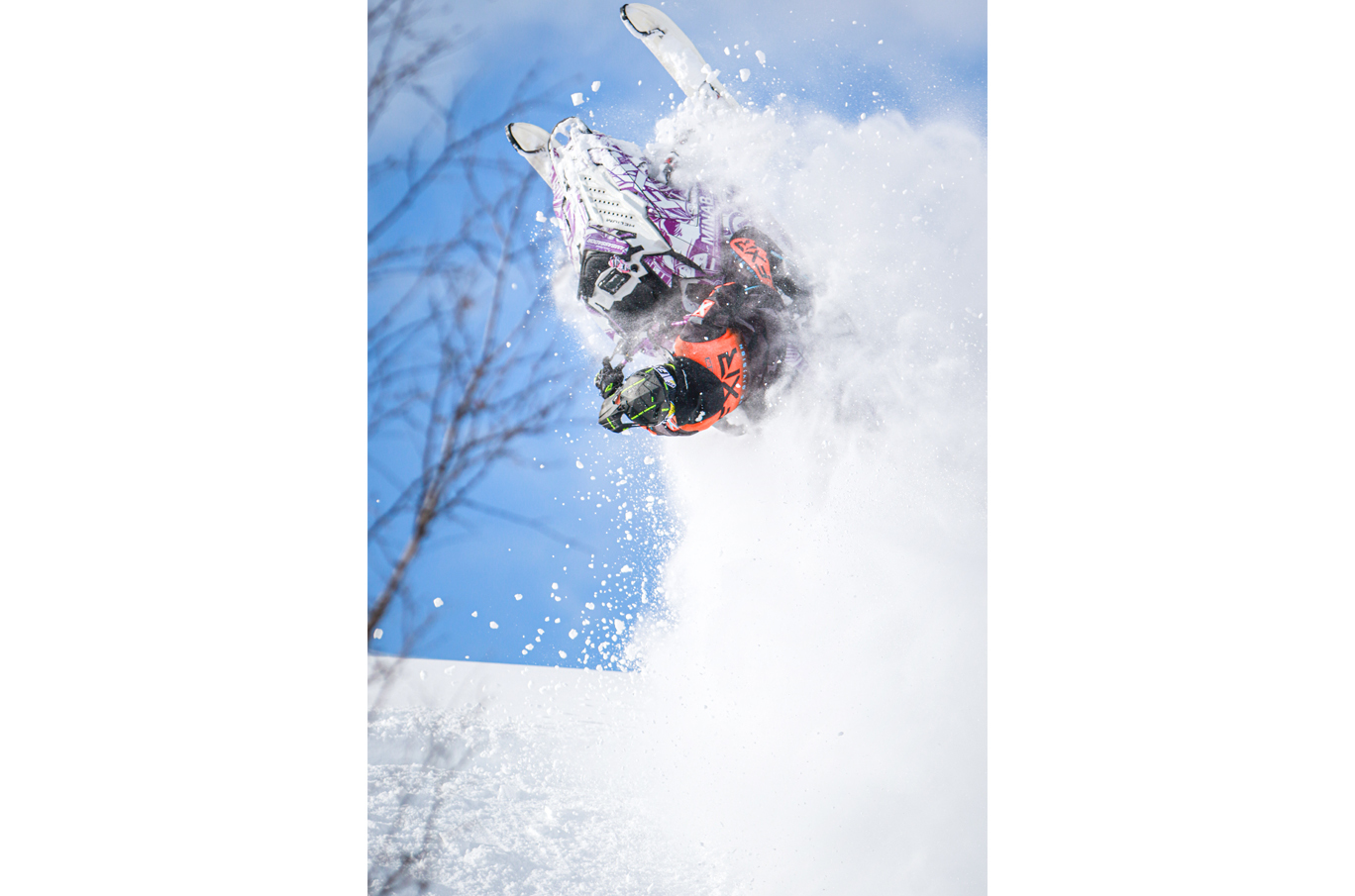 Snøscooterfører i oransje og svart drakt utfører en akrobatisk salto i lufta, omgitt av snøsprut og blå himmel