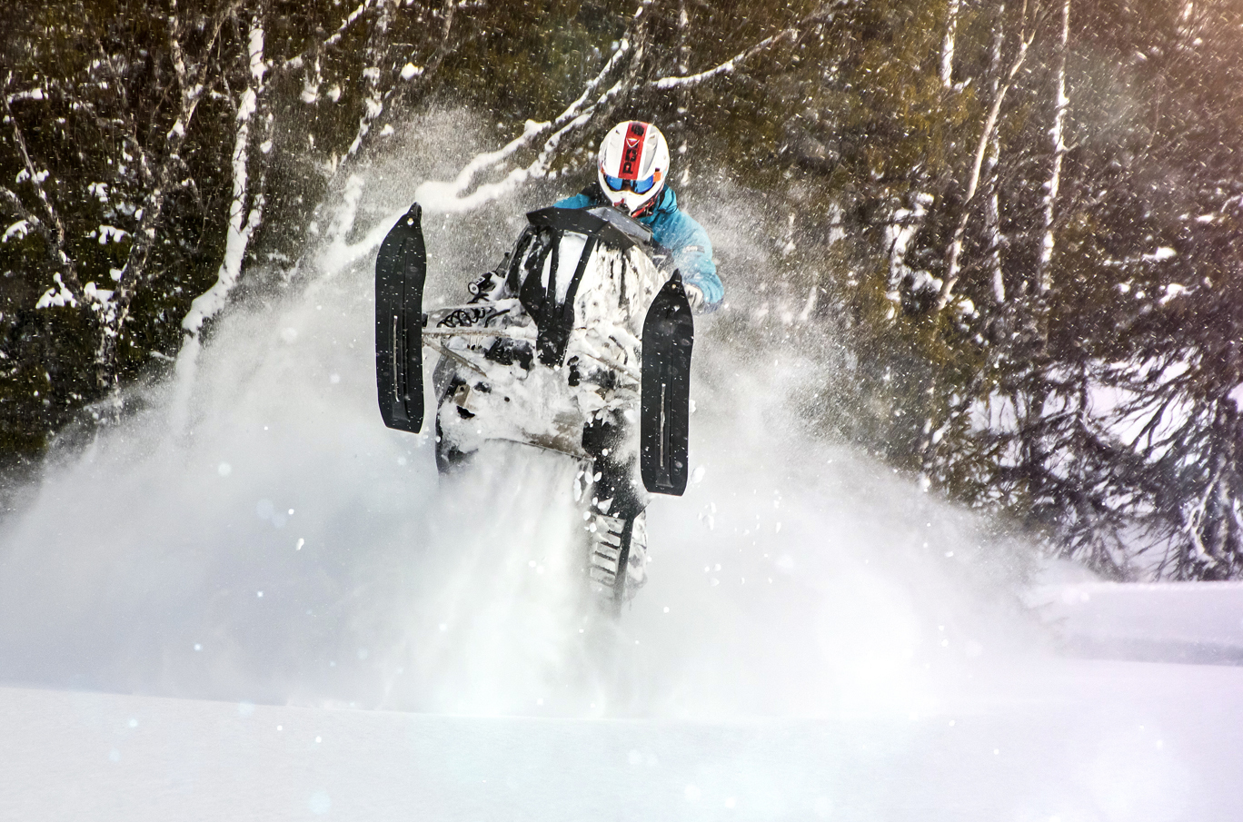 Snøscooter som hopper gjennom dyp snø i vinterskog, omgitt av virvlede snøflak og sollys