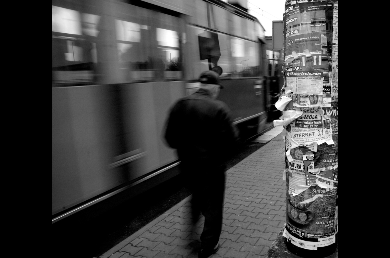 En mann med lue går langs fortauet ved en trikk som kjører forbi. Til høyre ses en stolpe dekket av reklameplakater.