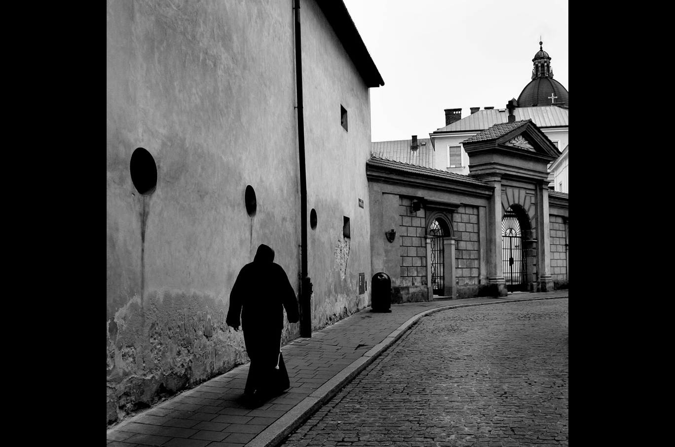 En person med hettegenser går langs en smal brosteinsgate ved en murvegg i svart-hvitt