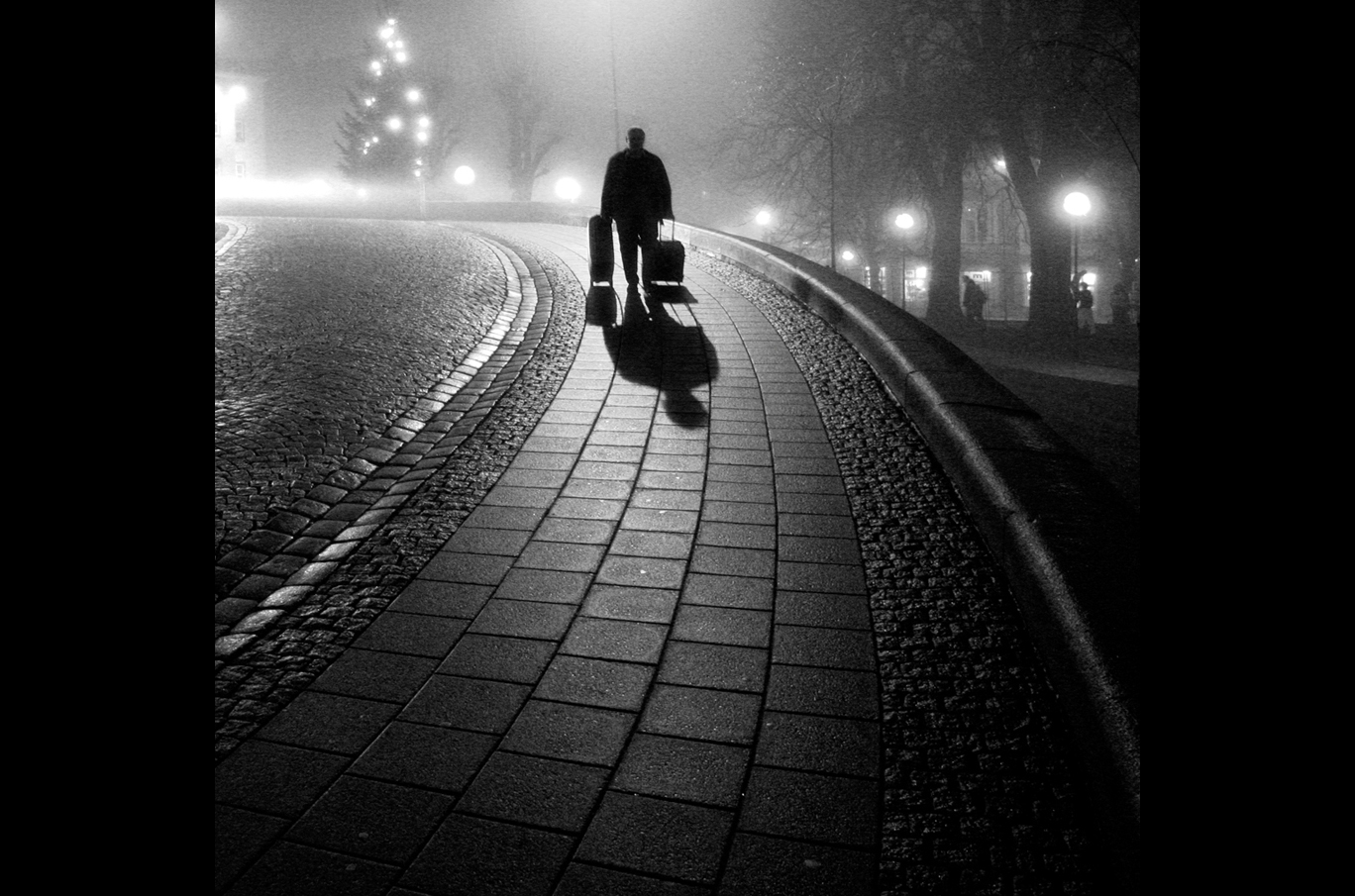En person går langs en svingete brosteinvei om natten, med to kofferter og lange skygger i lyset fra gatelykter