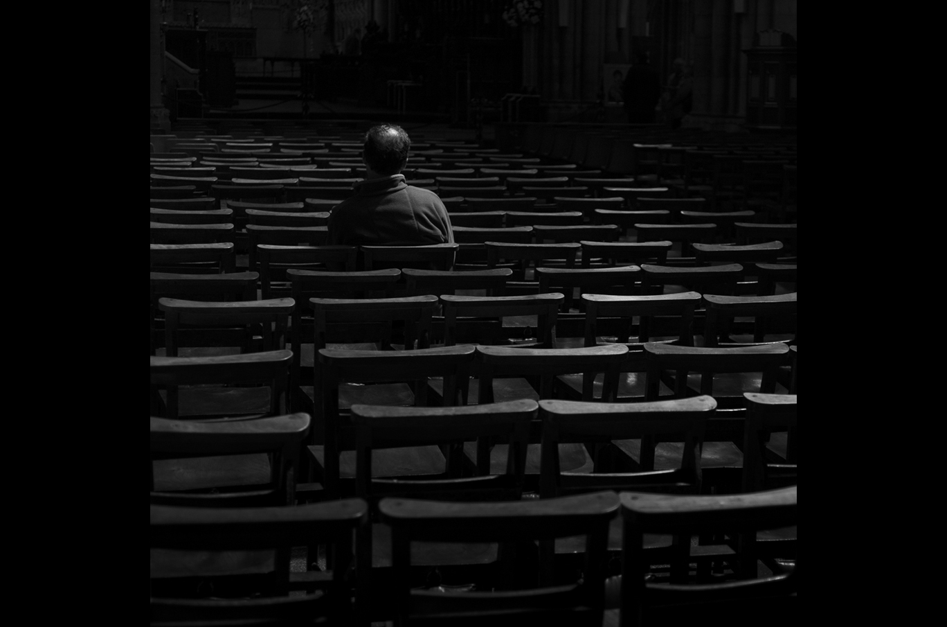En mann sitter alene midt i en rekke tomme trestoler i et mørkt kirkerom