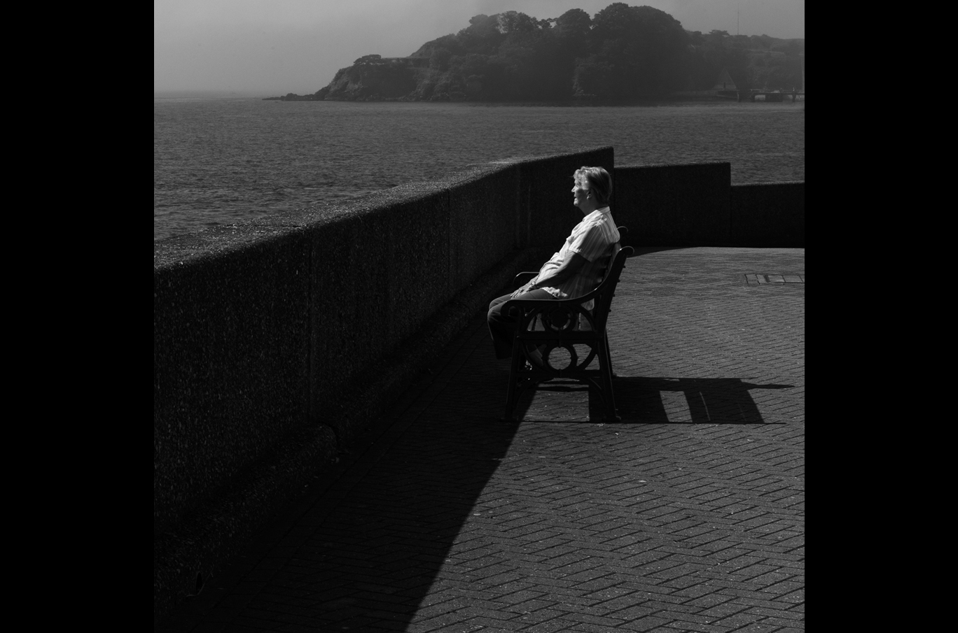 En mann sitter alene på en benk ved en steinmur, ser utover havet under grå, disige omgivelser
