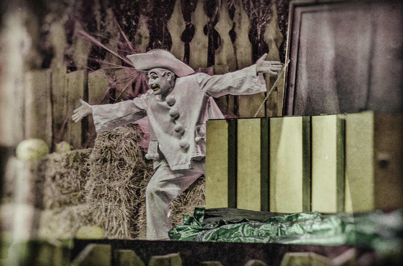 En person iført en hvit klovnedrakt med store knapper står midt på en scene dekorert med halmballer og treplater