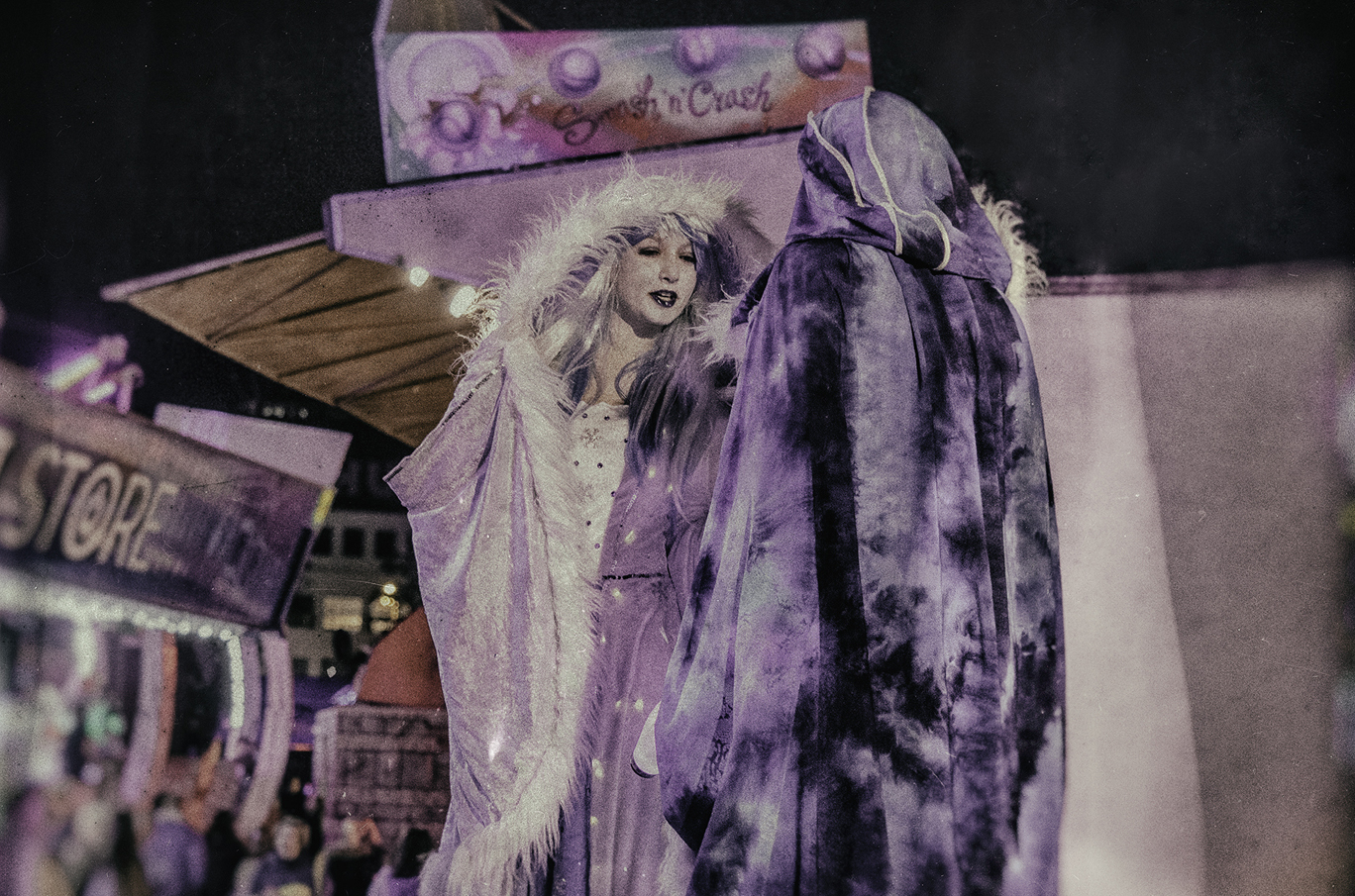 To personer i fantasifulle kostymer står foran en fargerik attraksjon på en nattlig festival.