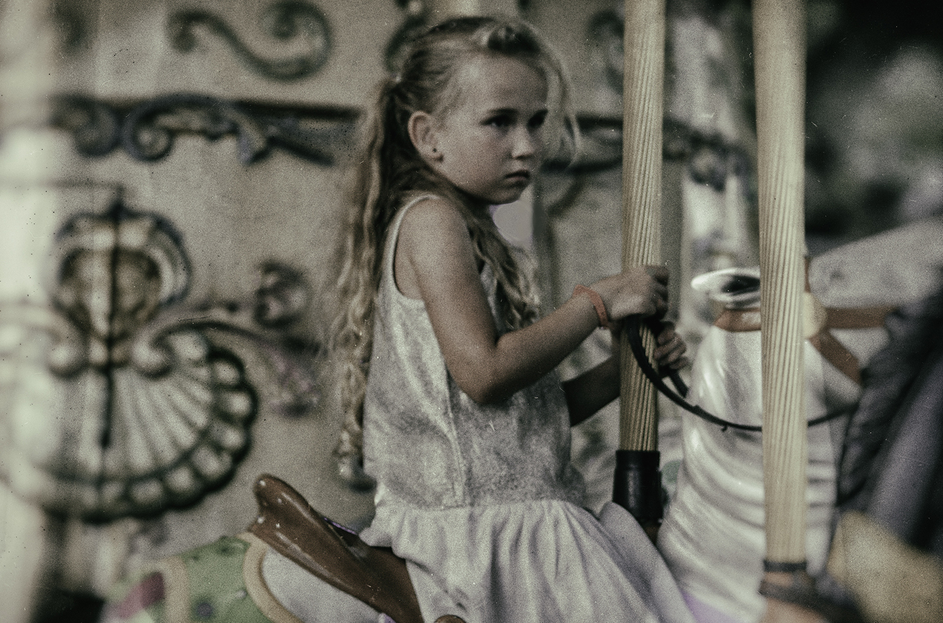 Et barn med langt, lyst hår sitter på en karusellhest og holder i tøylene. Bakgrunnen viser dekorative mønstre.