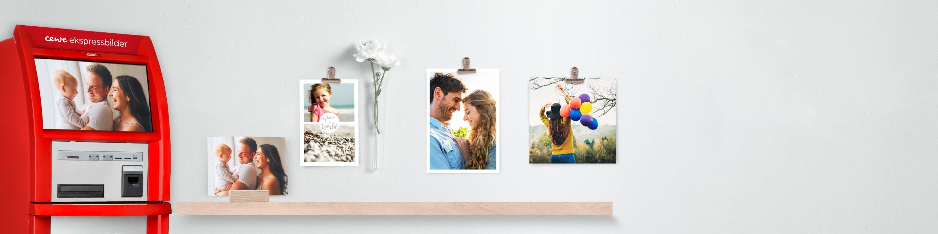 Rød CEWE-ekspressbilder automat ved siden av en hylle med utskrevne familiefotografier og en vase med hvite blomster