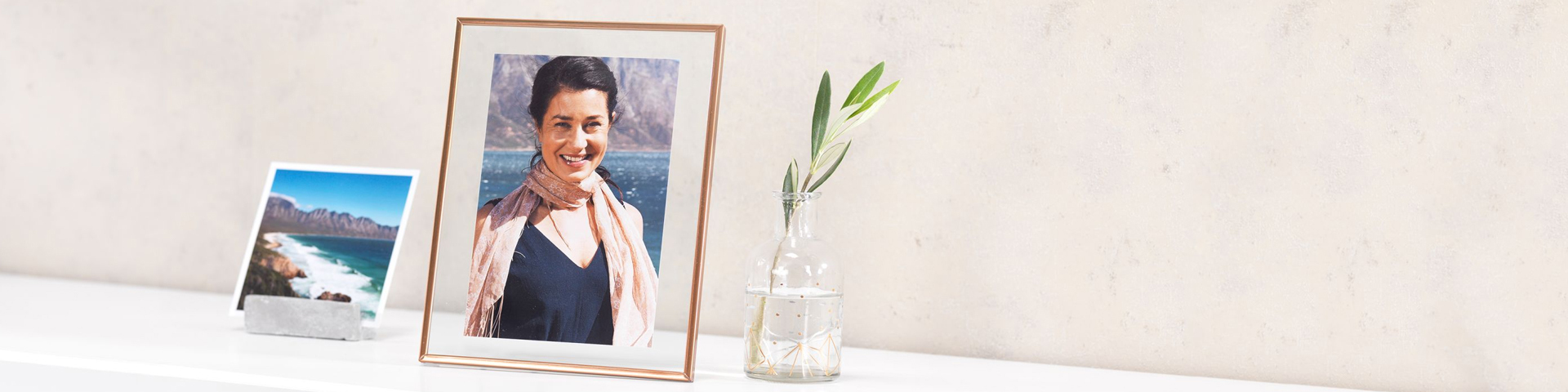 Innrammet fotografi med landskap i bakgrunnen, liten utskriftsstøtte med kystmotiv og glassflaske med olivengren.