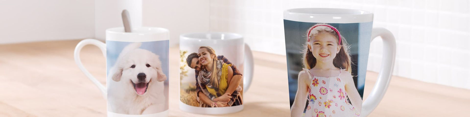 Tre kaffekopper med personlige bilder står på et lyst trebord; en kopp viser en valp, en viser et motiv av to klemmede personer, og en har et barn i sommerkjole