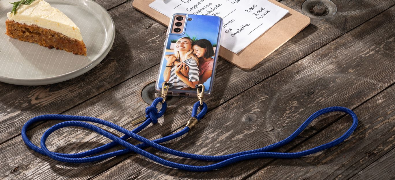 Mobiltelefon med personlig deksel og blå håndleddssnor, plassert på et rustikt trebord ved siden av en tallerken med gulrotkake og en meny.