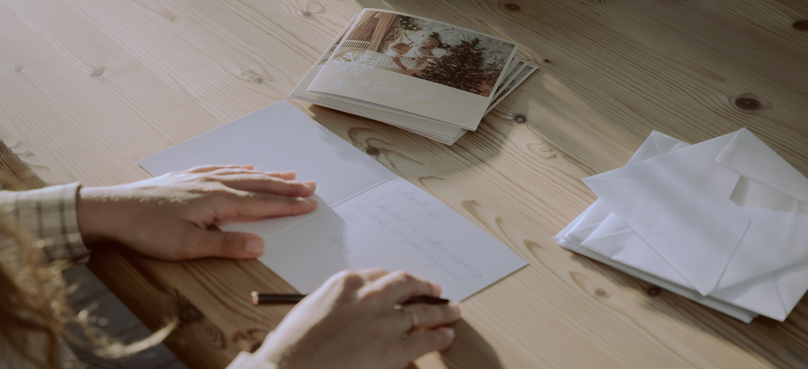 Hender som skriver en personlig julehilsen på et kort ved siden av en bunke ferdiglagde julekort og konvolutter