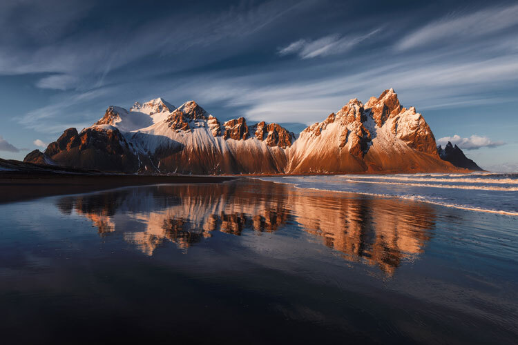 Snødekte fjell med oransje toner speiler seg i rolig vann under en blå himmel med lette skyer Snødekte fjell med oransje toner speiler seg i rolig vann under en blå himmel med lette skyer