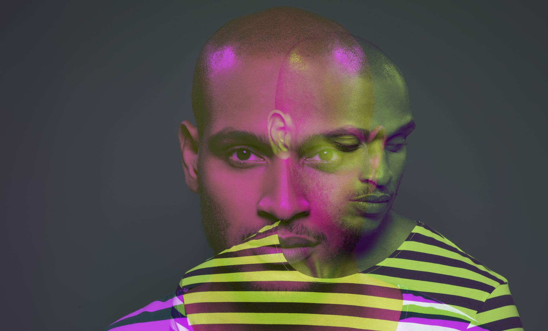 En mann med barbert hode iført en stripete skjorte i gule og lilla nyanser, belyst av fargerike neonlys