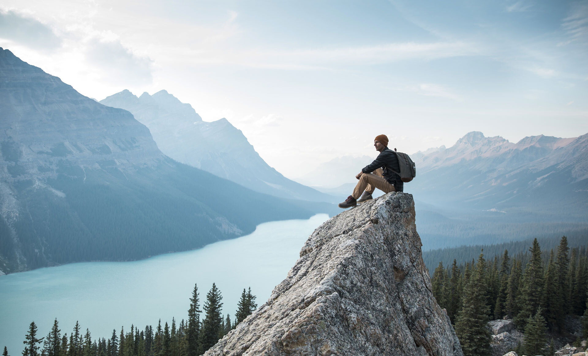 Person som sitter på en fjellklippe og ser utover en innsjø omgitt av fjell og skog