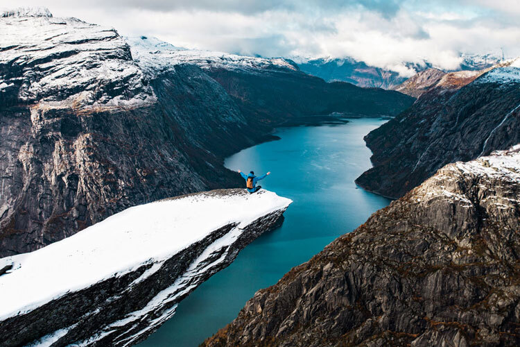 En person sitter på Trolltunga og ser utover en blå innsjø omgitt av snødekte fjell En person sitter på Trolltunga og ser utover en blå innsjø omgitt av snødekte fjell