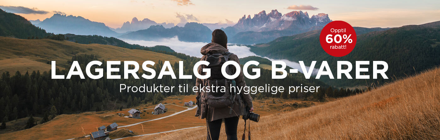 En person med ryggsekk og kamera står i en høstlig eng og ser utover et fjellandskap med hytter og skog