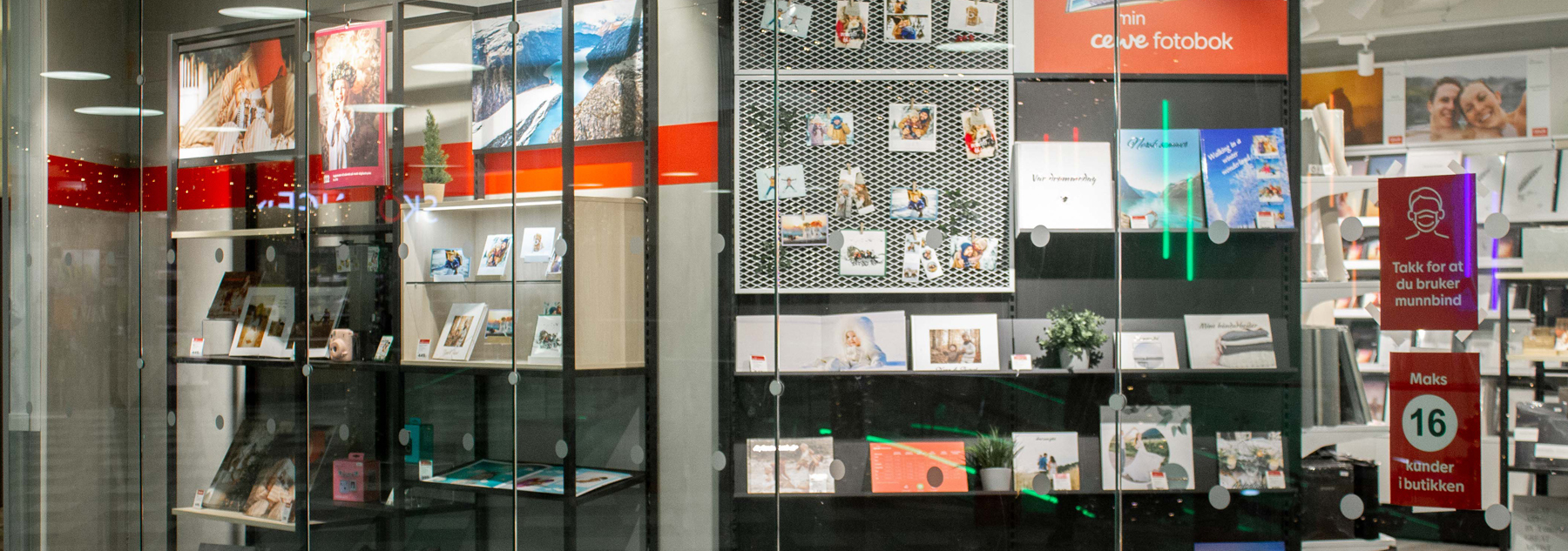 Utstillingsvindu med fotobøker, bilderammer og dekorative trykk på hyller, omgitt av glassflater og røde detaljer