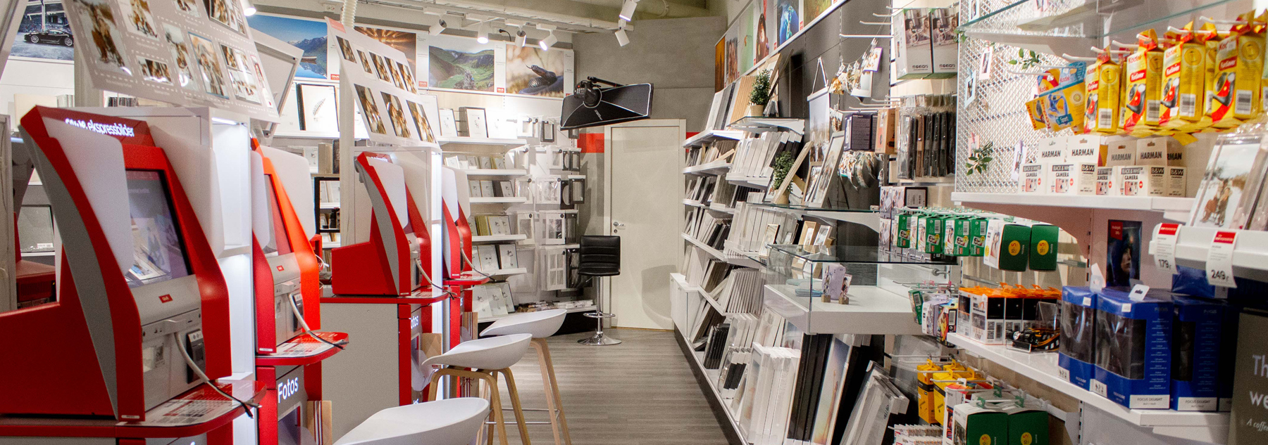 Lyse og moderne butikklokaler med fotokiosker i rødt og hvitt på venstre side, og hyller med rammer, fotoprodukter og tilbehør på høyre side
