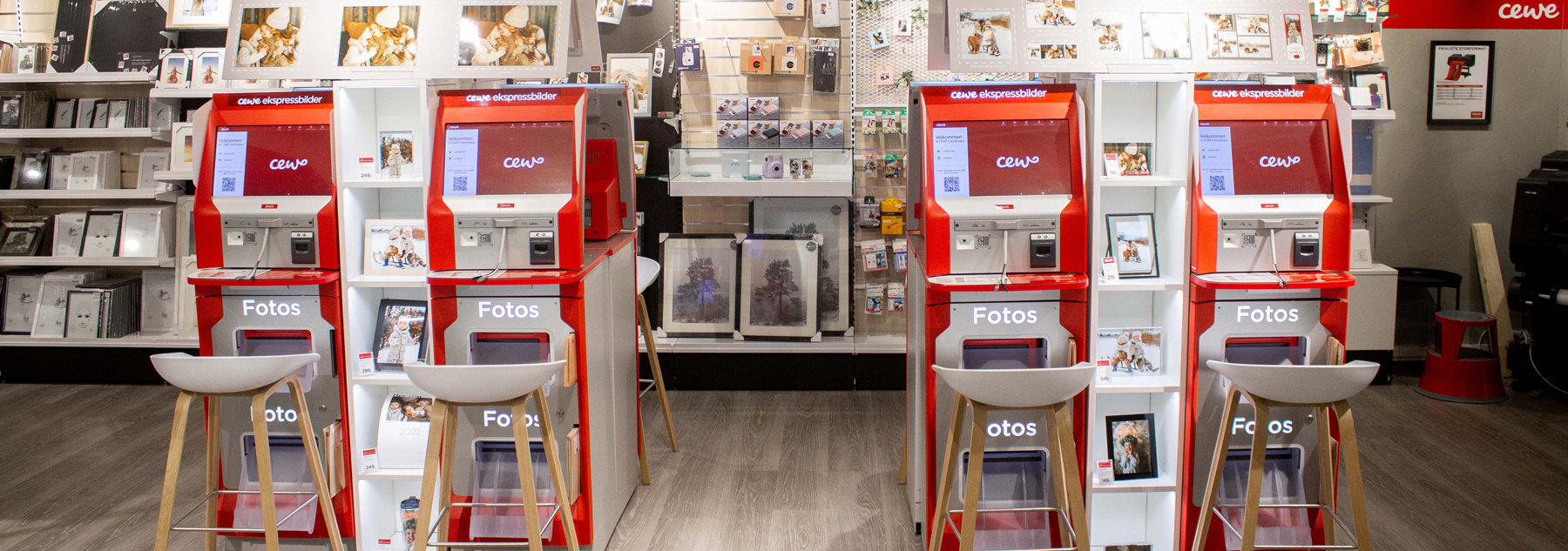 To røde CEWE-fotokiosker med skjermer og knapper, omringet av hvite hyller fylt med fotorammer, album og dekorasjoner i en butikk.