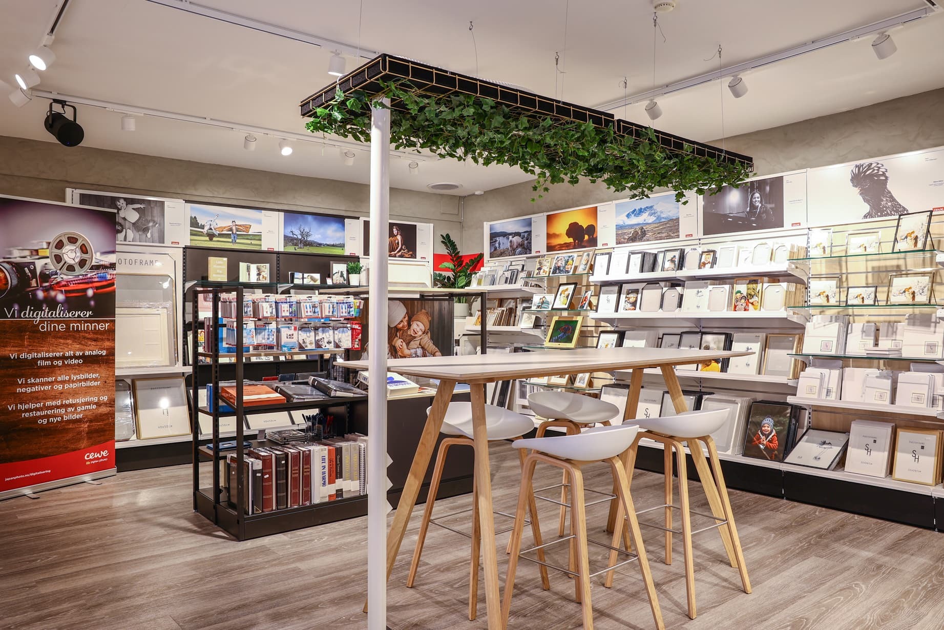 Lyst og innbydende butikklokale med moderne interiør, høye trebord med tilhørende stoler og hyller fylt med fotoprodukter og rammer