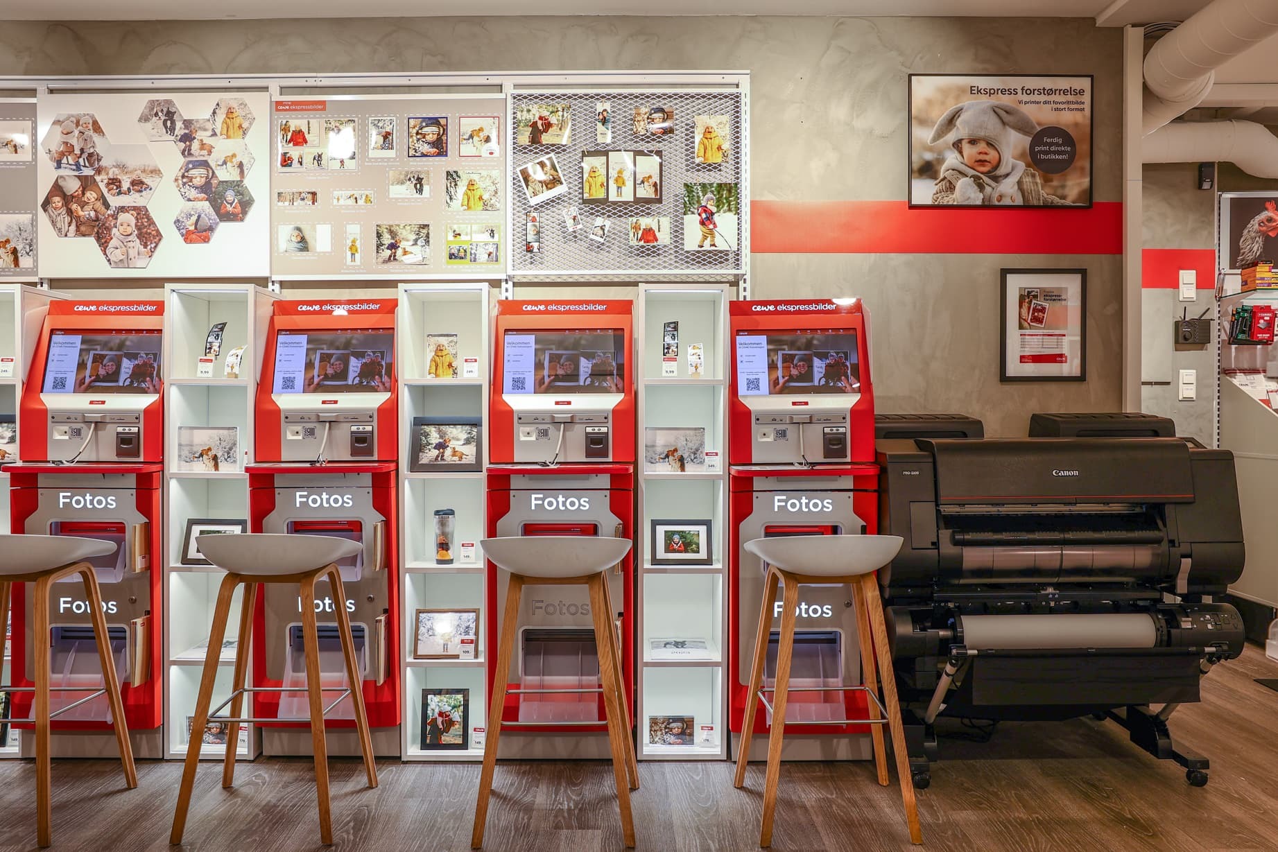 Røde CEWE fotokiosker med tre stoler foran, omgitt av veggmonterte bilder og en stor fotoskriver til høyre
