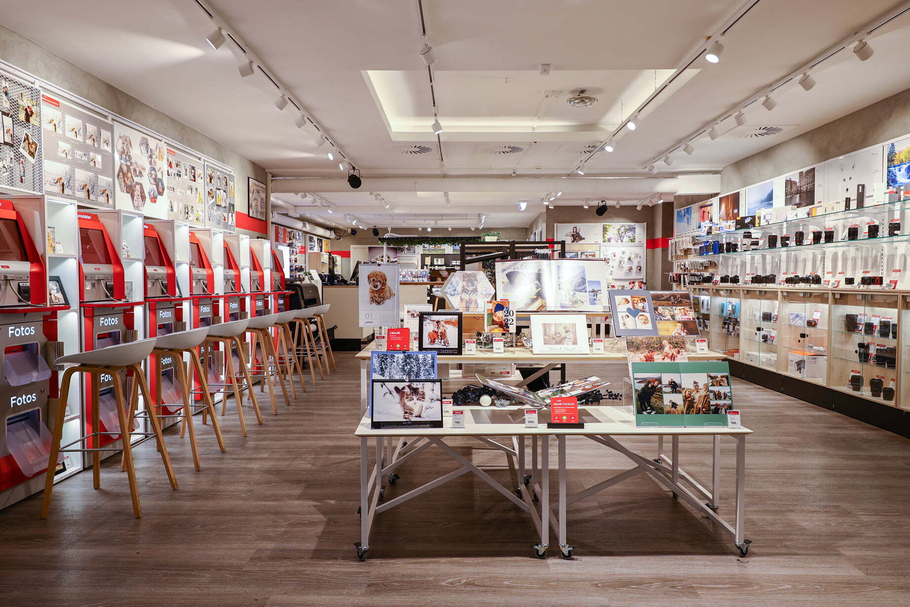 En lys og vennlig fotobutikk med moderne fotoautomater til venstre, høye stoler og utstilte fotoprodukter på bord
