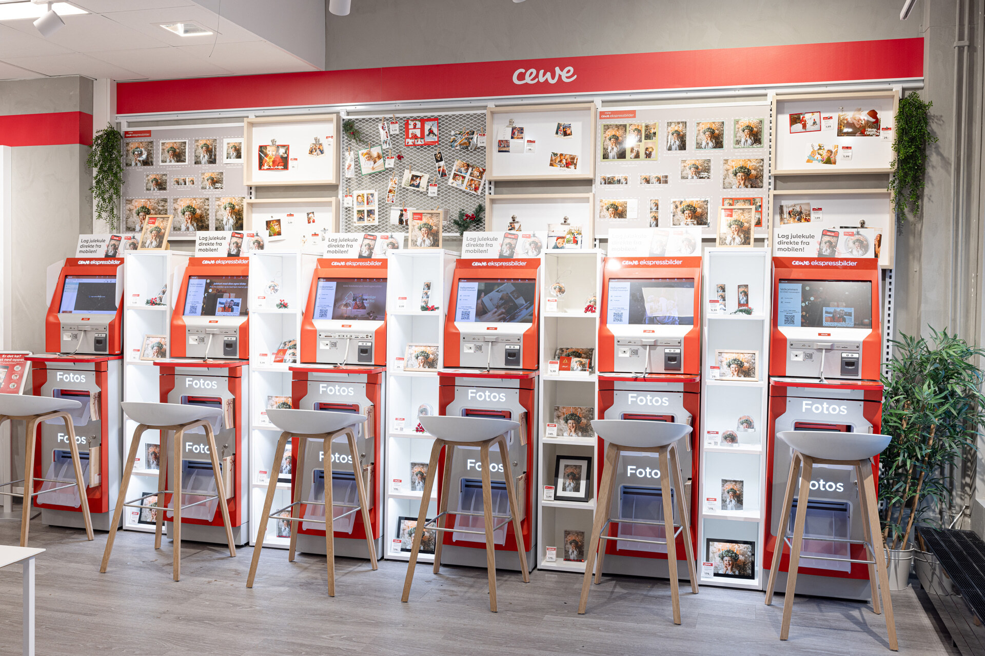 Røde CEWE fotokiosker på rad, omgitt av hyller med fotoeksempler og dekorasjoner, lysekrakker foran. Røde CEWE fotokiosker på rad, omgitt av hyller med fotoeksempler og dekorasjoner, lysekrakker foran.