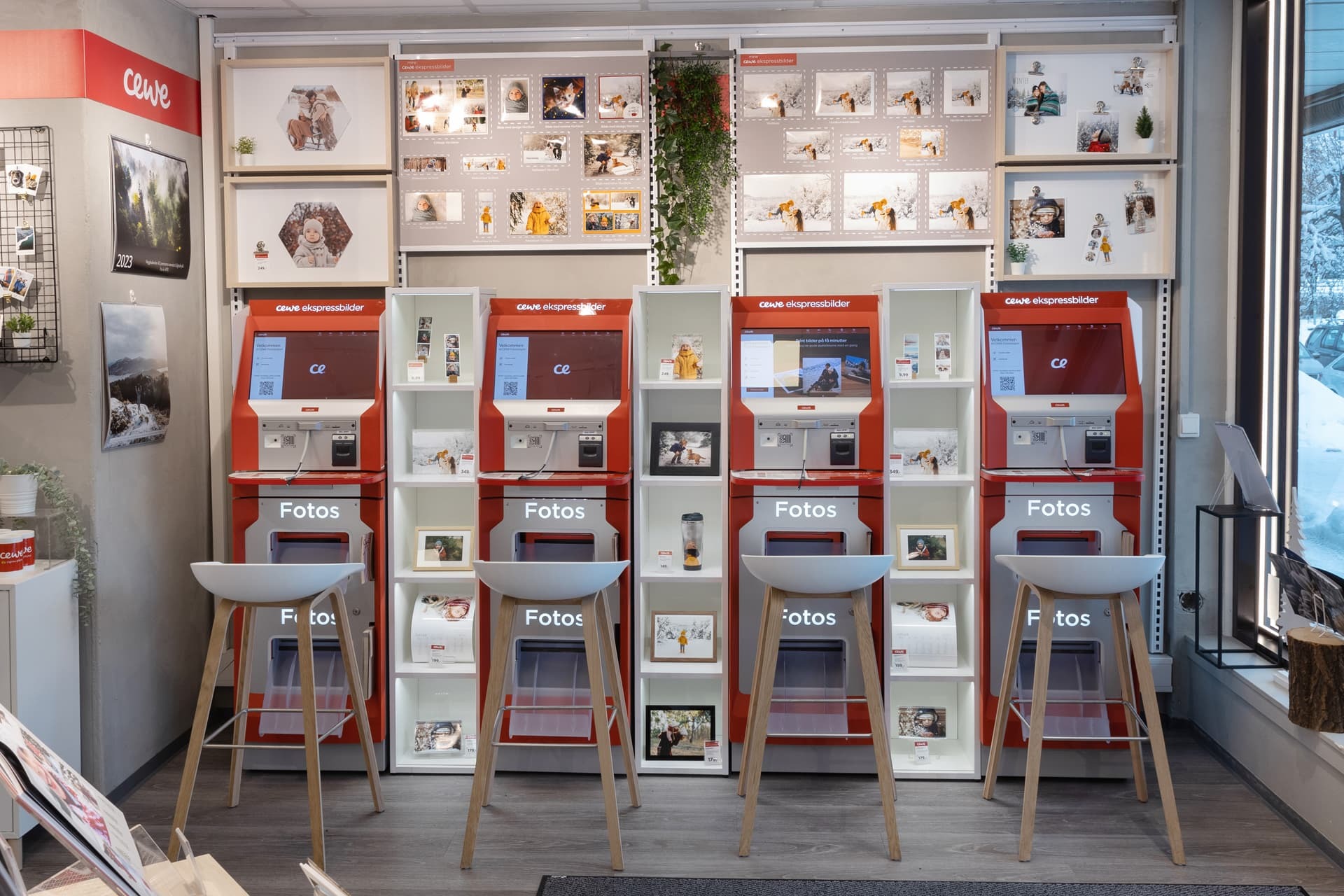 Fire røde CEWE-trykkskjermstasjoner med hvite hyller, utstilt i en butikk med fotoprodukter og dekorasjoner