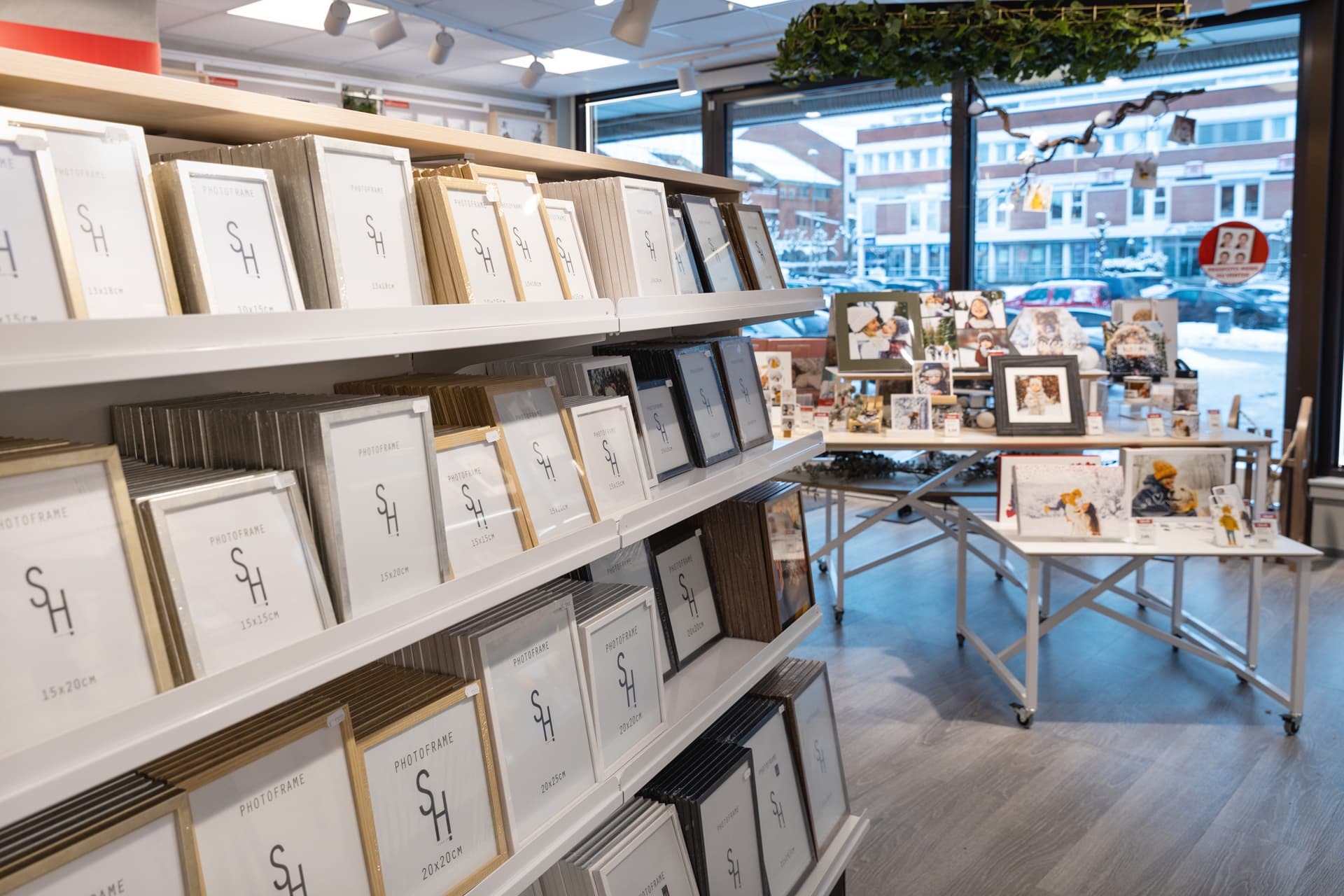 Hyller fylt med ulike bilderammer i gull, sølv, svart og hvit, samt bord med personlige fotogaver i et lyst butikklokale