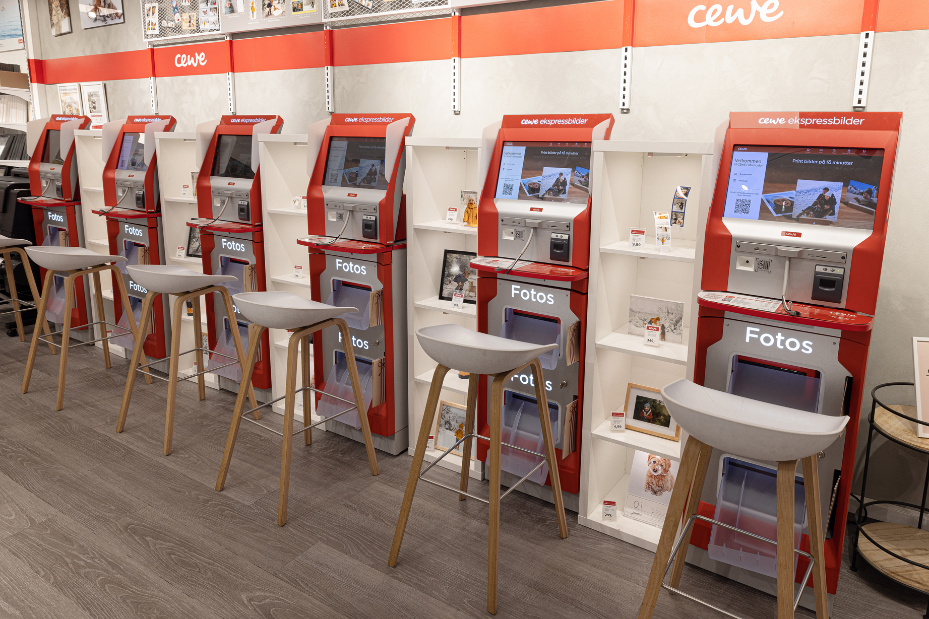 Røde CEWE fotokiosker med skjermer, stoler foran og hyller med fotoprodukter i bakgrunnen