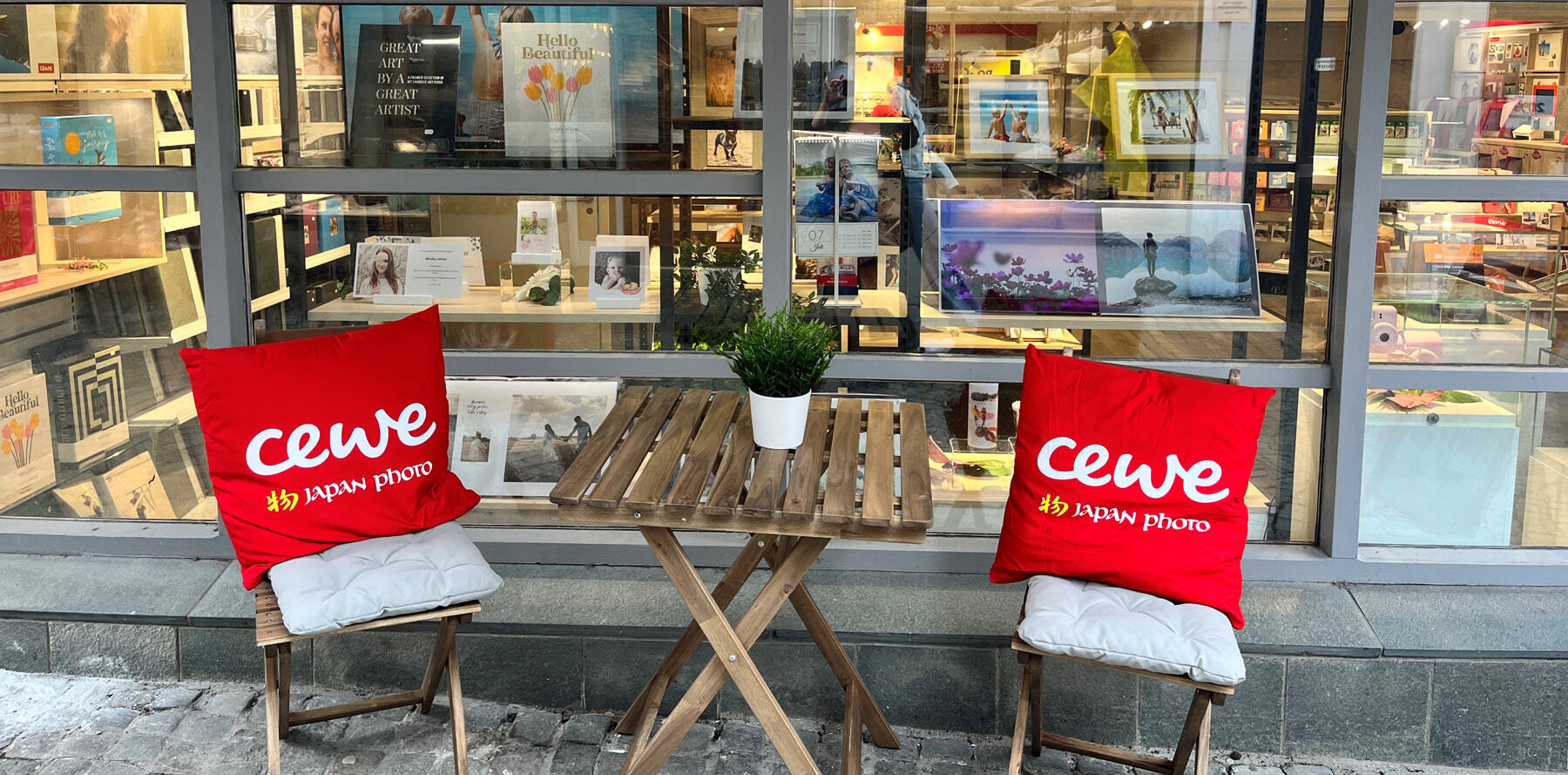 To utendørs stoler med røde CEWE Japan Photo-puter og et trebord med en grønn plante foran et butikkvindu som viser fotoprodukter.