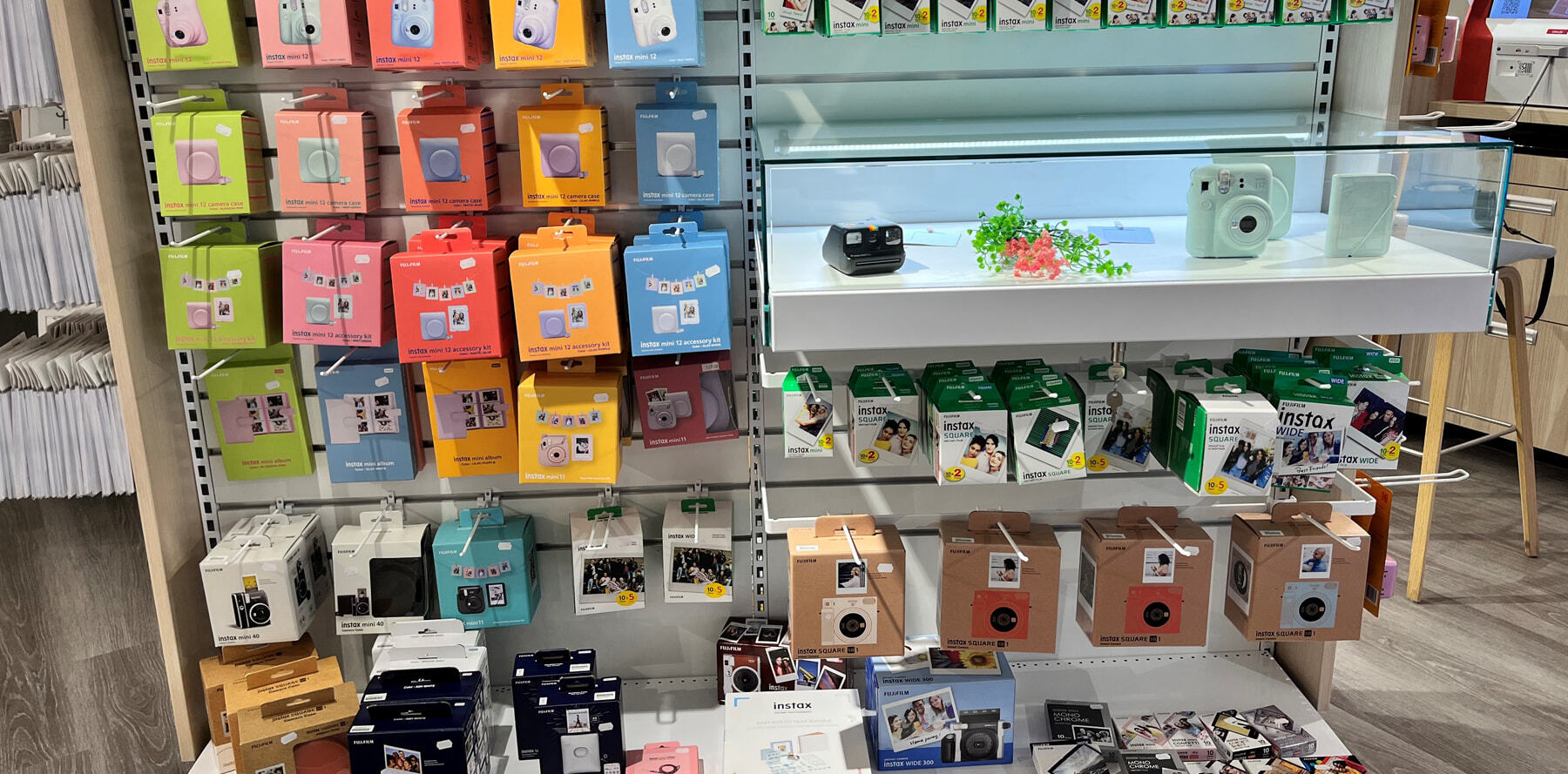 Hyller med fargerike Instax-produkter, inkludert kameraer, film og tilbehør i ulike størrelser og varianter