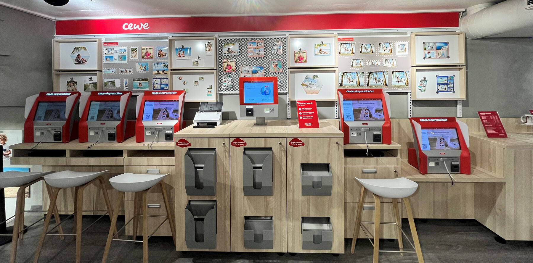 Fem røde CEWE-skriverstasjoner på lys trereduksjon, omgitt av utstilte bilder, rammer og sommermotiver