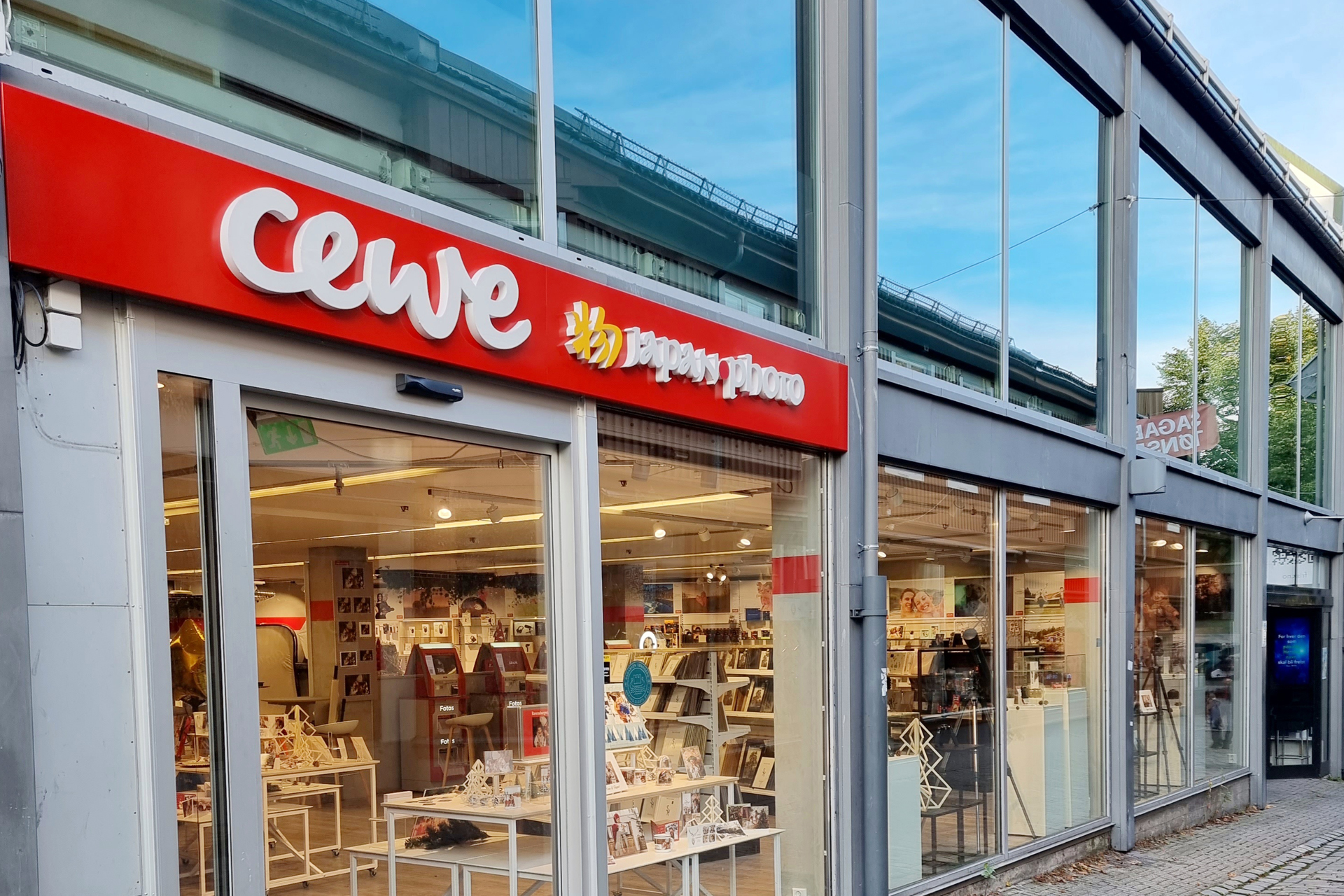 Butikklokale med CEWE Japan Photo-logo på rød bakgrunn, store vinduer som viser fotoprodukter og innredning inni butikken.