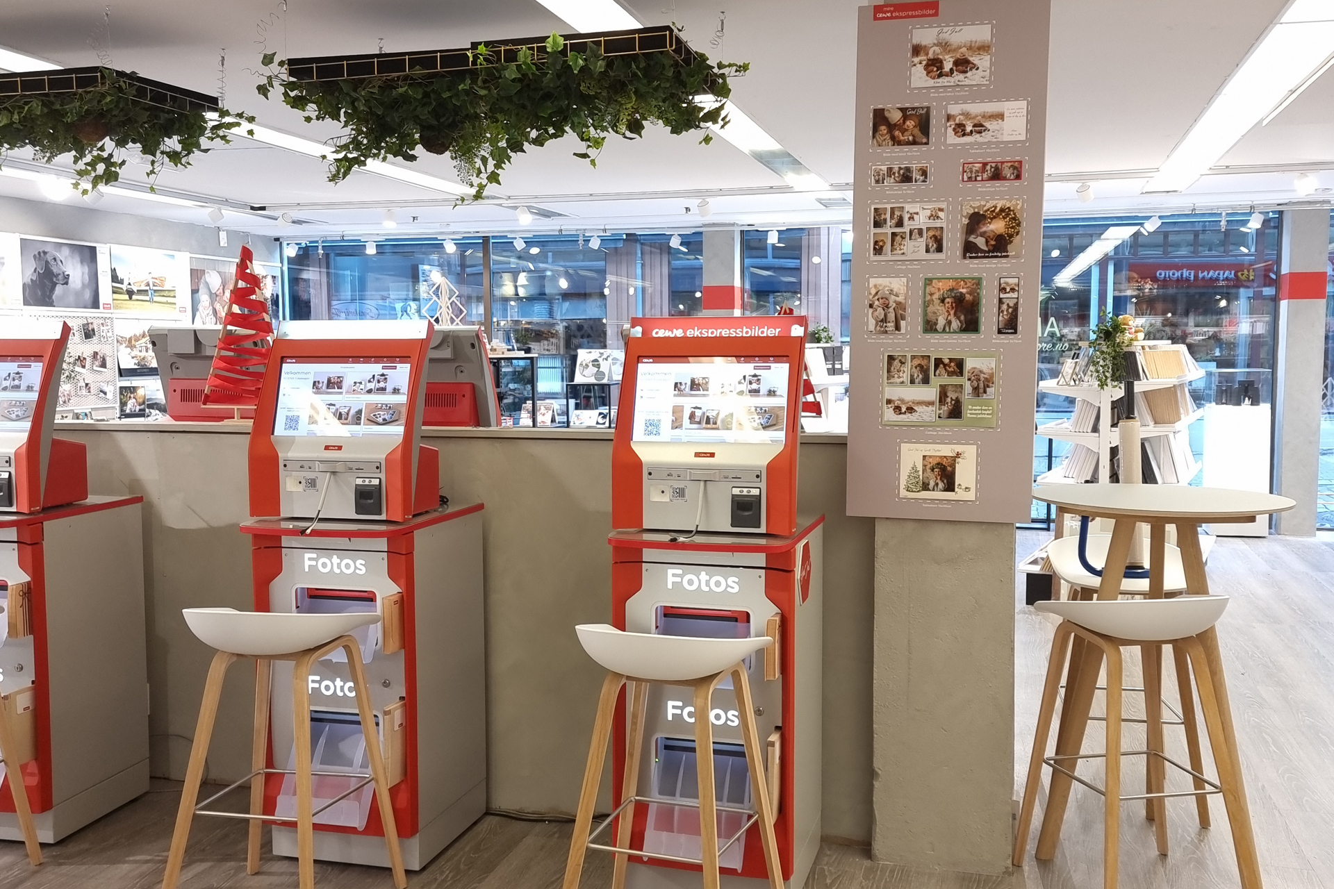 Tre fotostasjoner i lysegrå og rød design med skjermer for bildeutskrift. Grønne planter henger fra taket over.