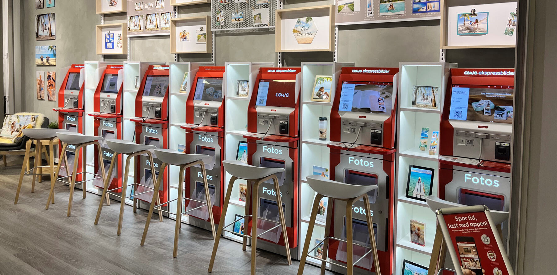 Røde CEWE fotokiosker på rad med høy barstoler foran, omgitt av inspirerende fotoutstillinger på veggen