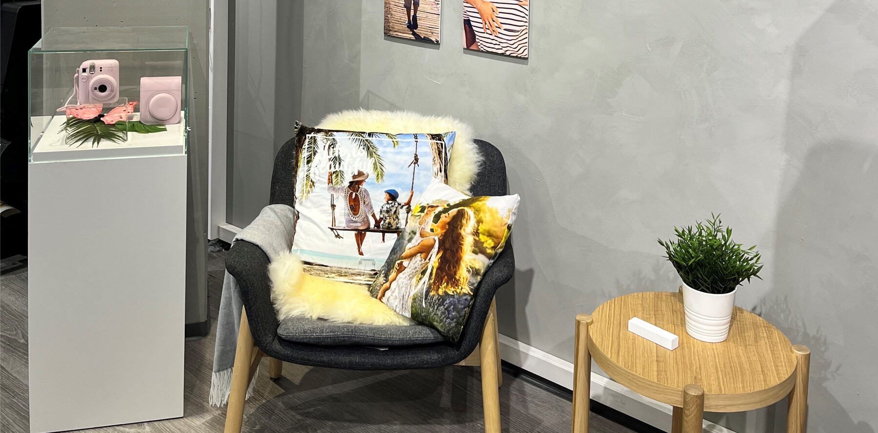 Grå stol med mykt pelspledd og to fargerike puter med fotomotiver av glade sommerminner, ved siden av liten trebord med hvit plantepotte