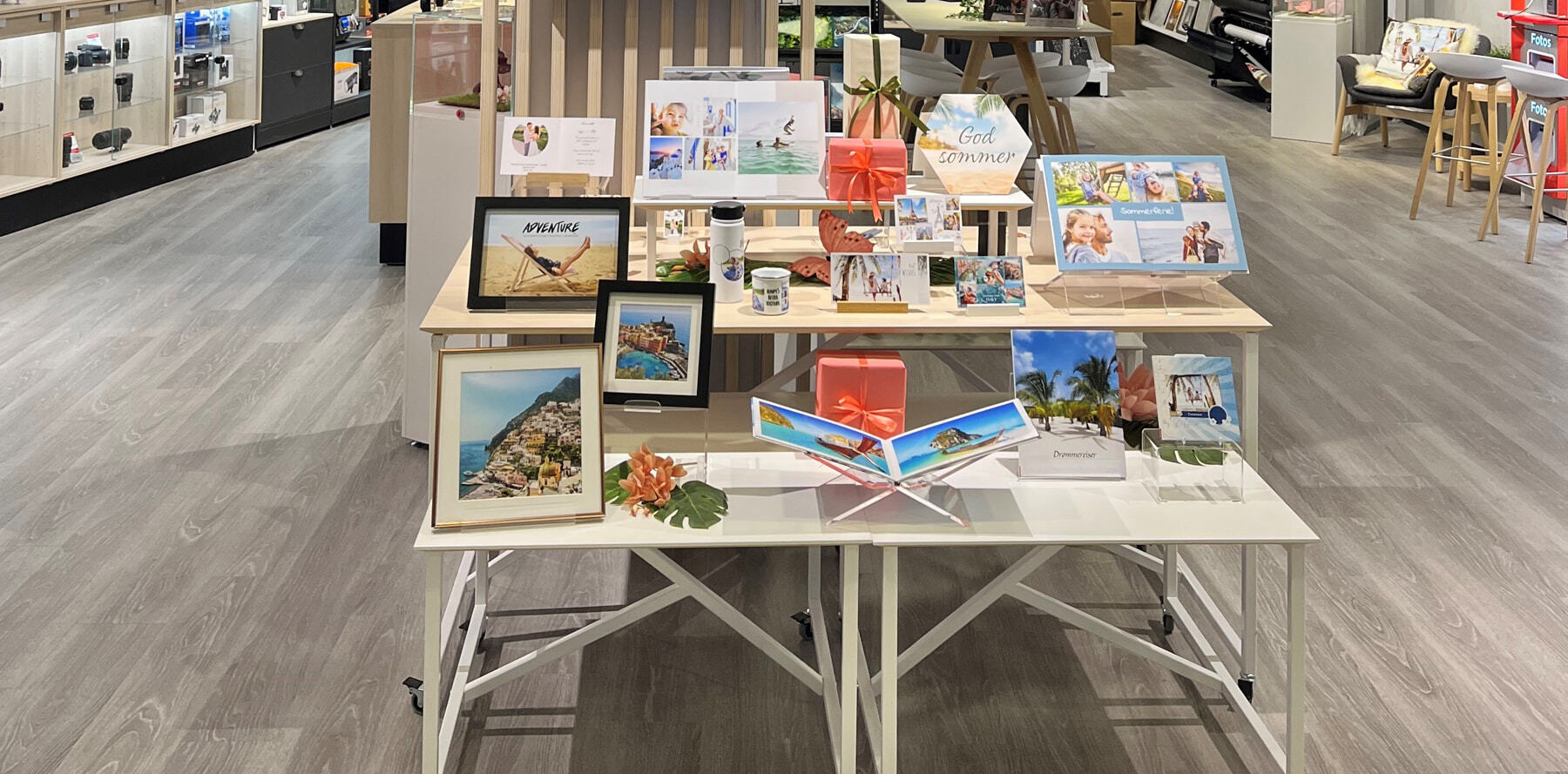 Utstillingsbord med fotoprodukter som fotobøker, bilderammer, krus og gaveløsninger, dekorert med sommerlige farger og blomster.