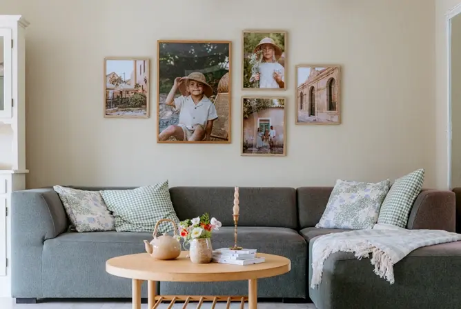 Grå sofa med grønne og blomstrete puter, rundt trebord med en vase med blomster, en tekanne og en lysestake, vegg dekorert med fem innrammede fotografier
